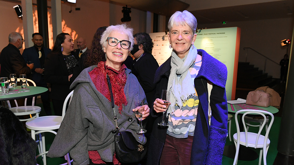 Two older women smile to camera, holding glasses of wine. People gather in groups behind them at an event. 