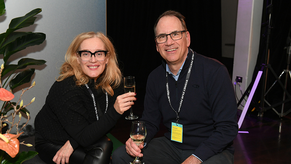 A woman and a man smile to camera at an event. They are both wearing glasses and holding glasses of wine. 
